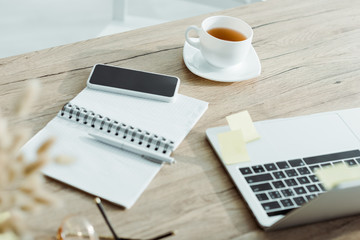 selective focus of laptop near smartphone with blank screen, cup of tea and notebook