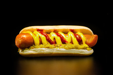 Classic American fast food: juicy hot dog with big pork sausage, tomato ketchup, cheese sauce and sauce with pickles and chili pepper. Black background, close-up.