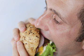  Close-up. A hungry young man eats a big hamburger sandwich with beef, tomatoes, onions, sauce, cheese, lettuce and a sesame bun on a light blue background. The concept of fast food.