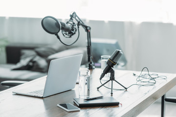 wooden desk with microphone, laptop, smartphone, notebook and glasses of water in broadcasting studio