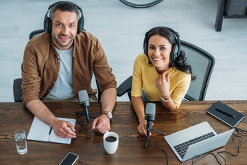 two cheerful radio hosts in headphones sitting at workplace and smiling at camera