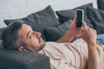 smiling man in earphones lying on sofa and holding smartphone with blank screen