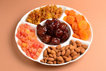 Mix of dried fruits and nuts on a white plate. Apricot, almond, raisin, dates fruit. On a brown background. Space for text or design.