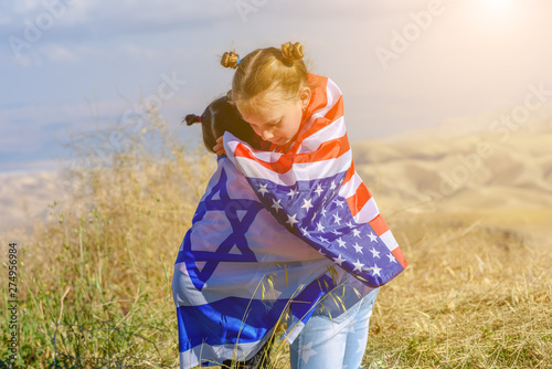 Two cute girls with American and Israel flags. Little children holding Israeli and USA flags hugging on meadow with beautiful landscape in background.Two nations one heart concept.
