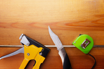 A staple gun, pocket knife, and a measuring tape are laying on a wooden surface