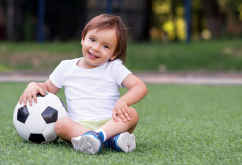 Little toddler boy sitting with legs crossed on football field in summer day with soccer ball. Happy child playing football outdoors. Active childhood concept