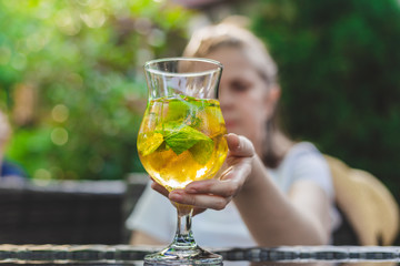Young woman sitting at a terrace grabbing a glass with ice tea that is placed on the table – Sweet summer refreshment with lemon slices and mint served on hot days