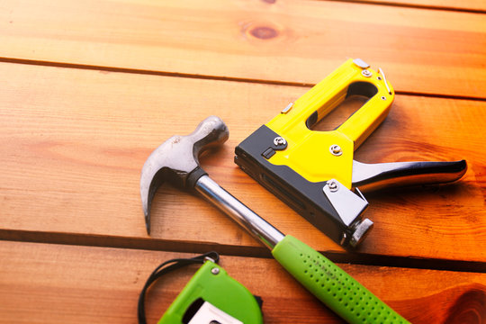 A hammer, measuring tape, and a staple gun are laying on a wooden surface