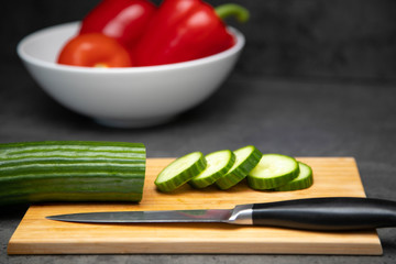 sliced fresh cucumber on a cutting board with a knife and a ceramic plate of vegetables.