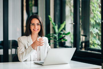 Portrait of elegant young female leader sitting at desk with laptop and holding a mug.