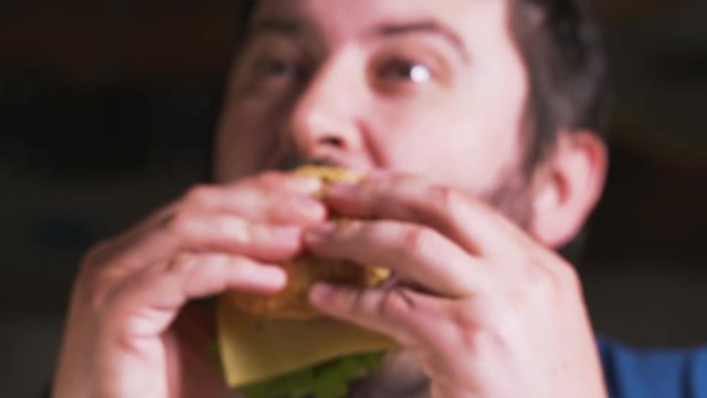 Happy smiling adalt man with beard eating delicious burger meat with cheese and vegetables. Hungry young guy having stacked burger at cafe enjoys eating, chewing. Closeup Fast food restaurant concept