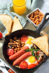 Breakfast in English scrambled eggs with bacon, beans, mushrooms, sausages, tomatoes and juice on light stone table.
