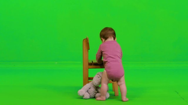 Adorable baby girl playing with her puppet while standing using a child's chair, from the back, over a green screen.