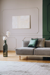White flowers in big green bottle shape like vase next to grey settee in fashionable living room interior