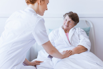 Senior grey woman lying in white hospital bed with young helpful nurse holding her hand
