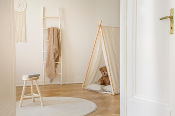 Tent with teddy bear in white scandinavian play room with wooden furniture and white carpet on the parquet, real photo with copy space