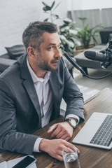 handsome radio host speaking in microphone while sitting at workplace near laptop