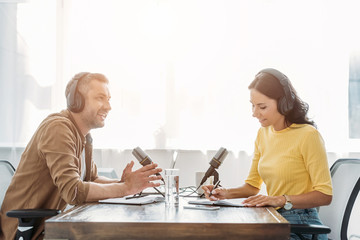 two smiling radio hosts in headphones talking while sitting at table in studio