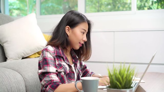 Freelance Asian woman working at home, business female working on laptop and using mobile phone talking with customer on sofa in living room at home. Lifestyle women working at home concept.