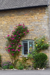 Fototapeta na wymiar Peasant house window with yellow and pink flowers. Stone facade and solid tiled roof of luxury medieval three bedroom cottage in the English countryside.