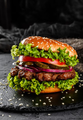 Beef burger with tomatoes, red onions, cucumber and lettuce on black slate over dark background. Unhealthy food - obrazy, fototapety, plakaty