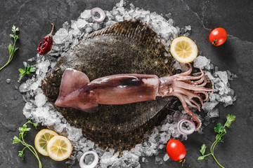 Raw whole flounder fish with whole squids on ice over dark stone background with lemon and spices....