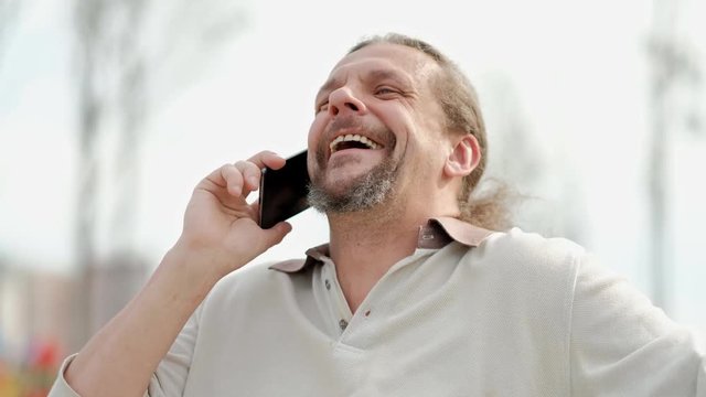 Relaxed attractive middle aged man with long gray hair speaks on smartphone, laughs and smiles. Strong positive emotions.