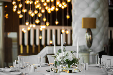 Wedding table setting is decorated with fresh flowers and white candles. Wedding floristry. Bouquet with roses, hydrangea and eustoma. On the background blur are burning garlands with light bulbs.