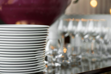 A stack of clean white plates stands on a table with a white tablecloth in an indoor restaurant. Buffet at the party. Blank empty glasses on the background