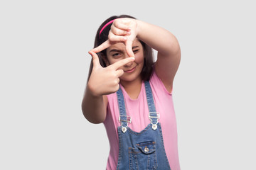 Portrait of attentive brunette young girl in casual style, pink t-shirt and blue denim overalls standing and looking into crop hand composition gesture. studio shot, isolated on light gray background.