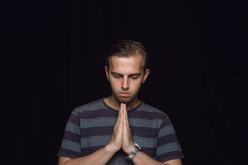 Close up portrait of young man isolated on black studio background. Photoshot of real emotions of male model. Praying with closed eyes, looks hopeful. Facial expression, human emotions concept.