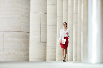 Full length portrait of beautiful business woman wearing red skirt walking towards camera along row of pillars, copy space