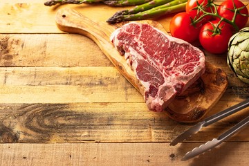 Fresh raw piece of marbled beef steak with fresh vegetables and tongs, on a cutting board, on a wooden background. Restaurant menu, home menu. Delicious food, gastronomy, culinary background