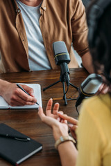 cropped view of radio host writing in notebook while interviewing woman in broadcasting studio