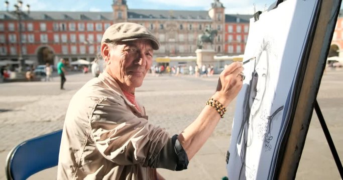 Portrait of happy elderly man painting in a public square in Madrid, Spain and looking at camera. Creative Spanish artist drawing with pencil on the street and smiling