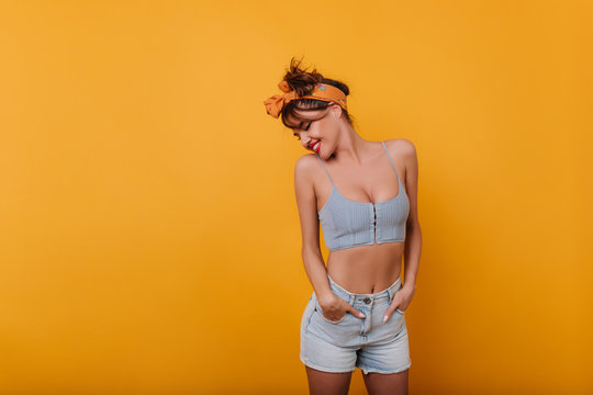 Adorable shapely girl with shy smile posing with hands in pockets. Indoor photo of blissful trendy european woman isolated on yellow background.