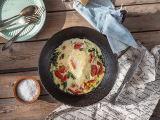 Omelet with tomatoes and spinach in a round cast iron pan on an old Board table, cotton napkins and coarse salt in a clay salt shaker, Cutlery, taken at close range