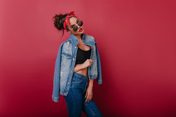 Serious fashionable girl in denim outfit standing in confident pose. Bored brunette woman with red ribbon in hair posing on claret background.