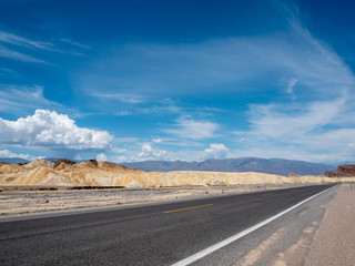 Fototapeta na wymiar Roadway and sedimentary rock hills in Death Valley National Park with brilliant blue sky and clouds.