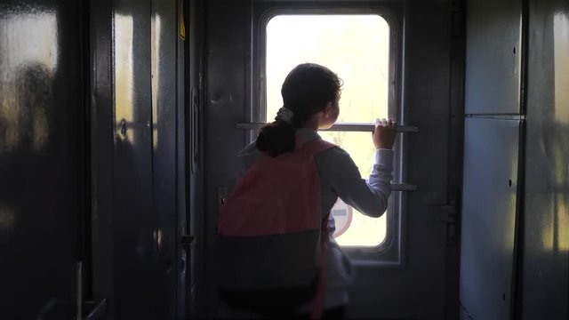 little teenage girl is a backpacker traveling by train. travel transportation railroad concept. tourist school girl in the train wagon lifestyle with backpack looking out the window waving his hand