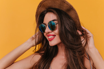 Close-up portrait of beautiful white woman with bright lipstick smiling to camera. Studio photo of relaxed caucasian girl in hat and black sunglasses.