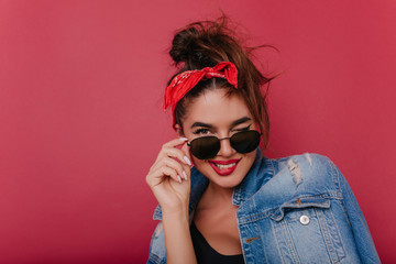 Close-up portrait of laughing happy girl with dark eyes. Attractive caucasian young woman touching her black sunglasses while posing on claret background.