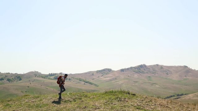 man with beard mustache with red backpack parachute green hat striped white yellow T-shirt jeans, walks through mountains summer and looks around. tattoo on hand. caucasian