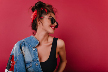 Beautiful brown-haired girl with black sunglasses laughing on claret background. Studio shot of cute smiling young woman holding denim jacket.