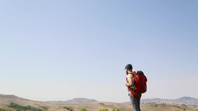 man with beard mustache with red backpack parachute green hat striped white yellow T-shirt jeans, walks through mountains summer and looks around. tattoo on hand. caucasian