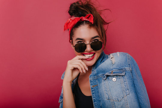 Close-up portrait of blissful lady in elegant sunglasses playfully posing on dark background. Studio photo of refined european girl with red ribbon having fun alone.