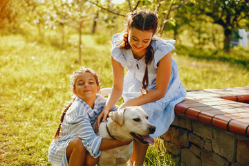 Fototapeta premium two beautiful and cute girls in blue dresses with beautiful hairstyles and make-up sitting in a sunny green garden and playing with a dog