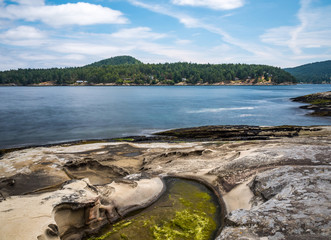 beautiful rocky shore line with green algae filled water puddle and green forest covered island over the horizon under cloudy blue sky