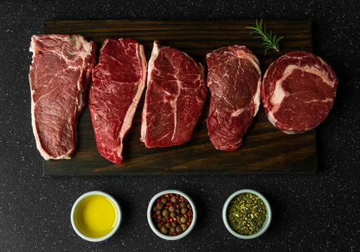 Selection of raw beef meat food steaks against black stone background. New york striploin steak, top blade, rib eye, and other cuts of meat.
