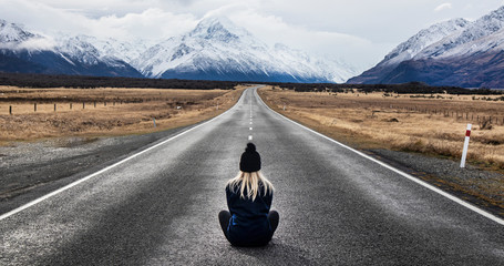 Girl with blonde hair wearing a hat sits in the middle of the road in New Zealand looking at the mountain view - Powered by Adobe